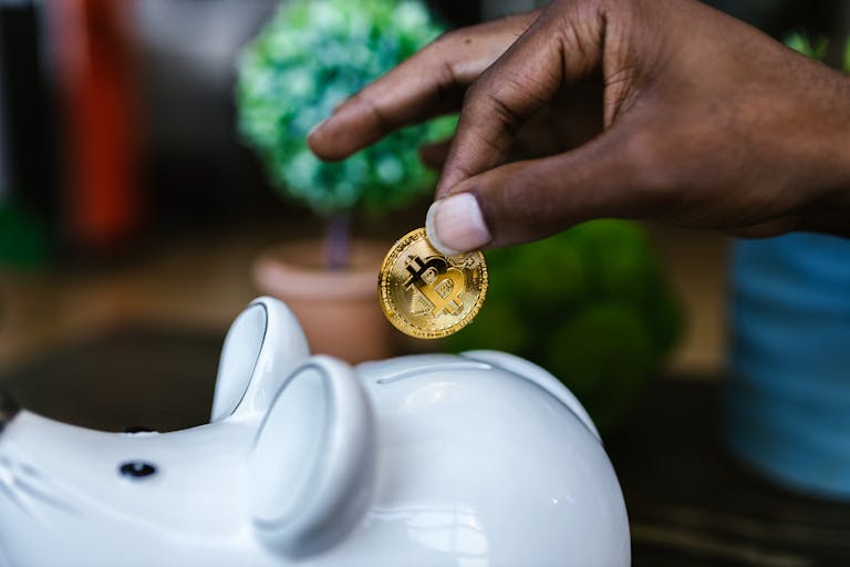 Juntos Seguros A close-up of a hand placing a bitcoin into a white piggy bank, symbolizing investment and savings.