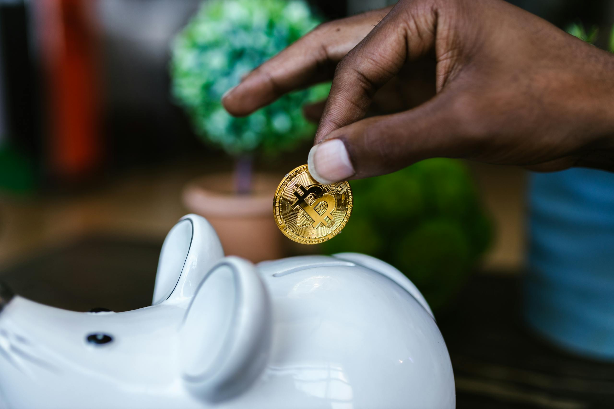 Juntos Seguros A close-up of a hand placing a bitcoin into a white piggy bank, symbolizing investment and savings.