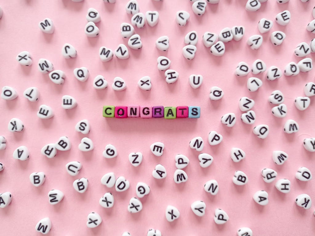 Shop Sunnylife Giant Jumbling Tower Colorful beads spell 'Congrats' on a pink backdrop with scattered letters.