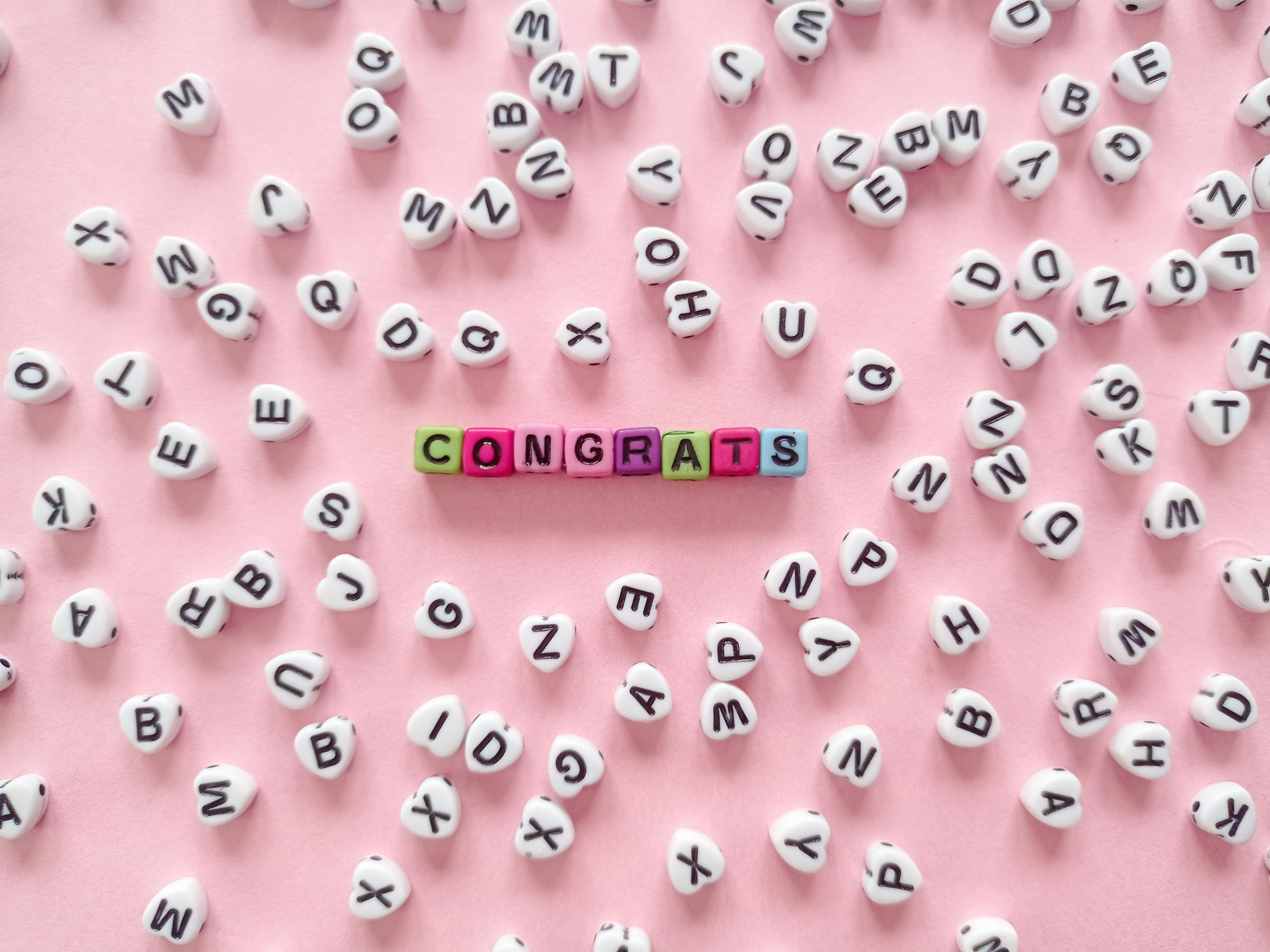 Shop Sunnylife Giant Jumbling Shop Sunnylife Giant Jumbling Tower Colorful beads spell 'Congrats' on a pink backdrop with scattered letters.
