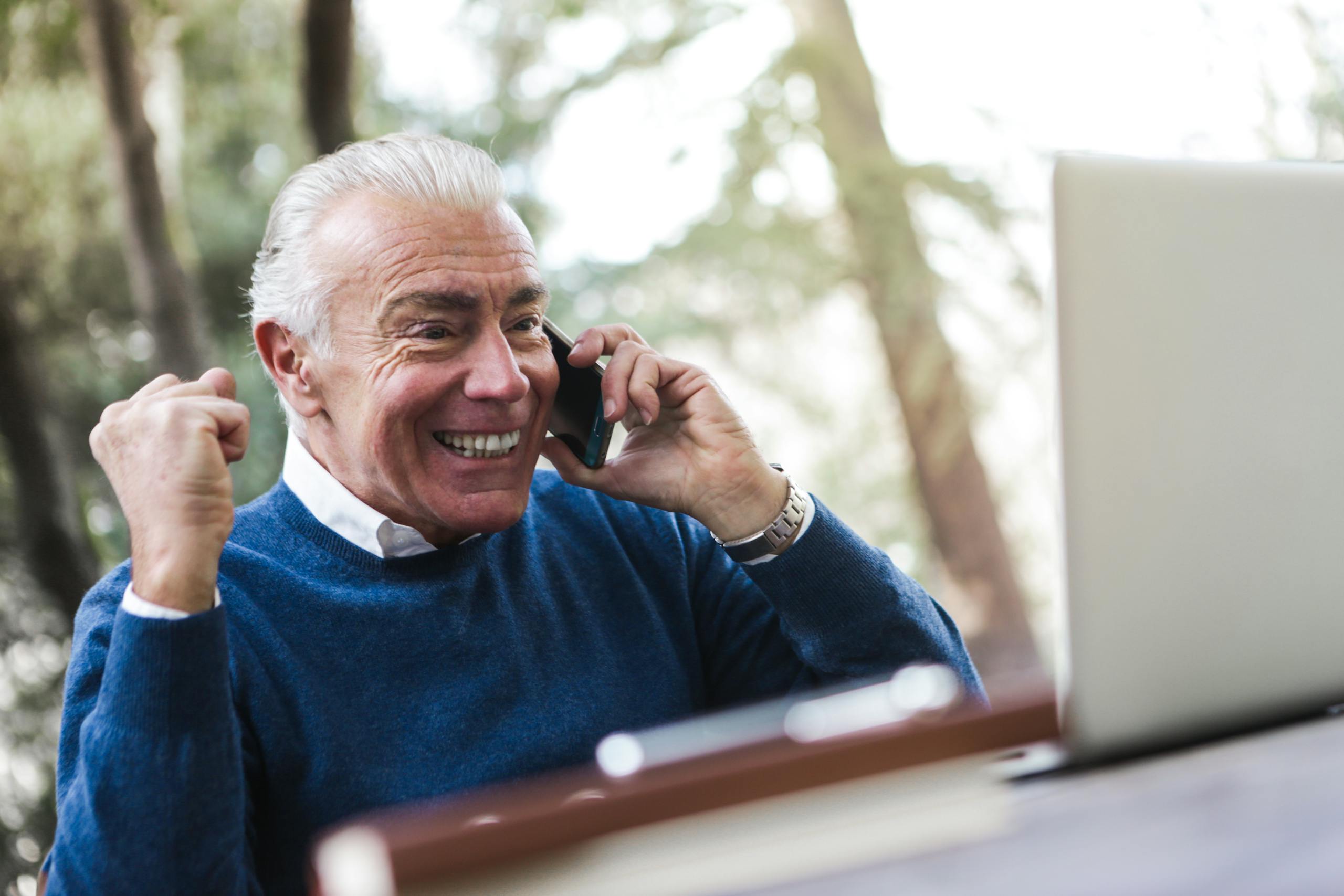 PermanentAd Elderly man smiling and cheering while talking on the phone using laptop outdoors.