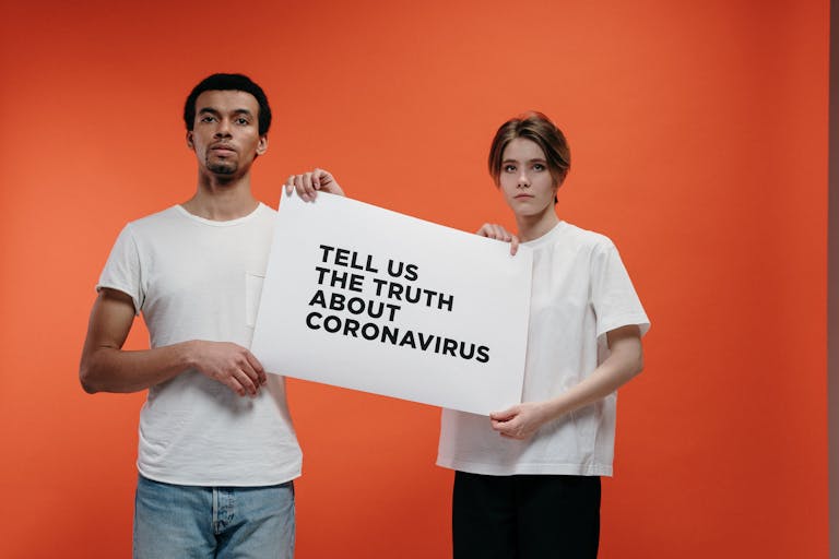 MC Lyte Car Crash Two people holding a 'Tell Us The Truth About Coronavirus' sign against a vibrant orange background.