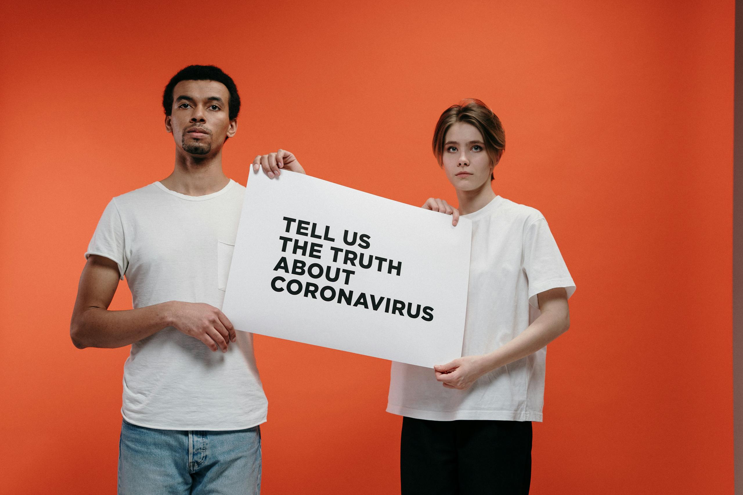 MC Lyte Car Crash Two people holding a 'Tell Us The Truth About Coronavirus' sign against a vibrant orange background.