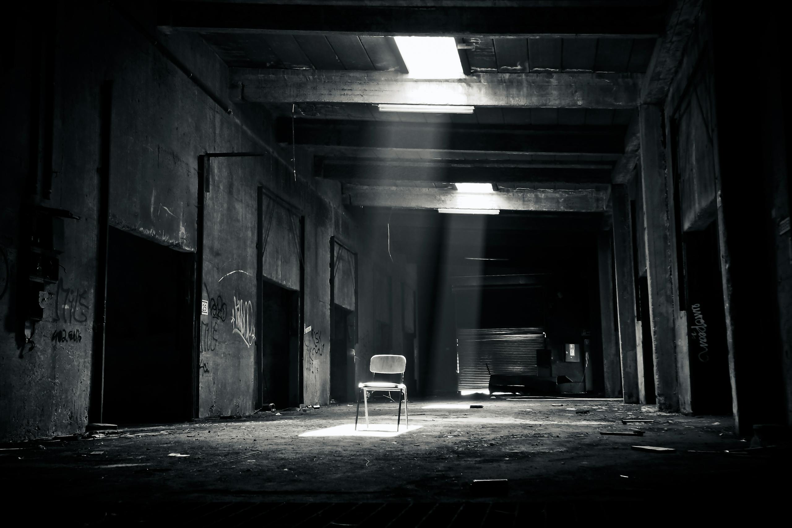 Carmela Clouth A haunting black and white photo of an abandoned warehouse with a solitary chair illuminated by sunlight through a window.