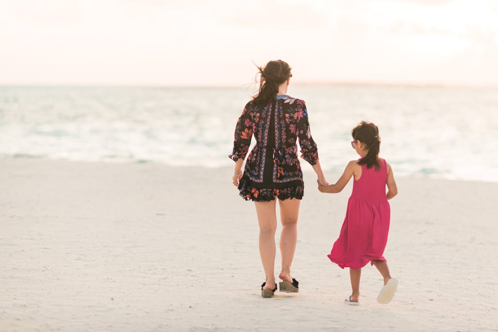 FamousParentingMomlife A heartwarming moment of a mother and daughter walking hand in hand on a tranquil beach.