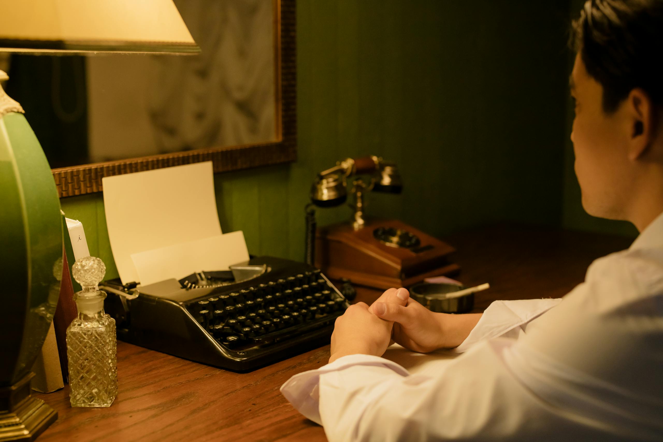 Serlig A man in a vintage setting sits at a desk with a typewriter and lamp, reflecting a nostalgic workplace.