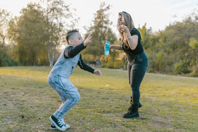 A mother and son FamousParentingMomlife enjoy blowing bubbles together in a sunny park, capturing a joyful family moment.