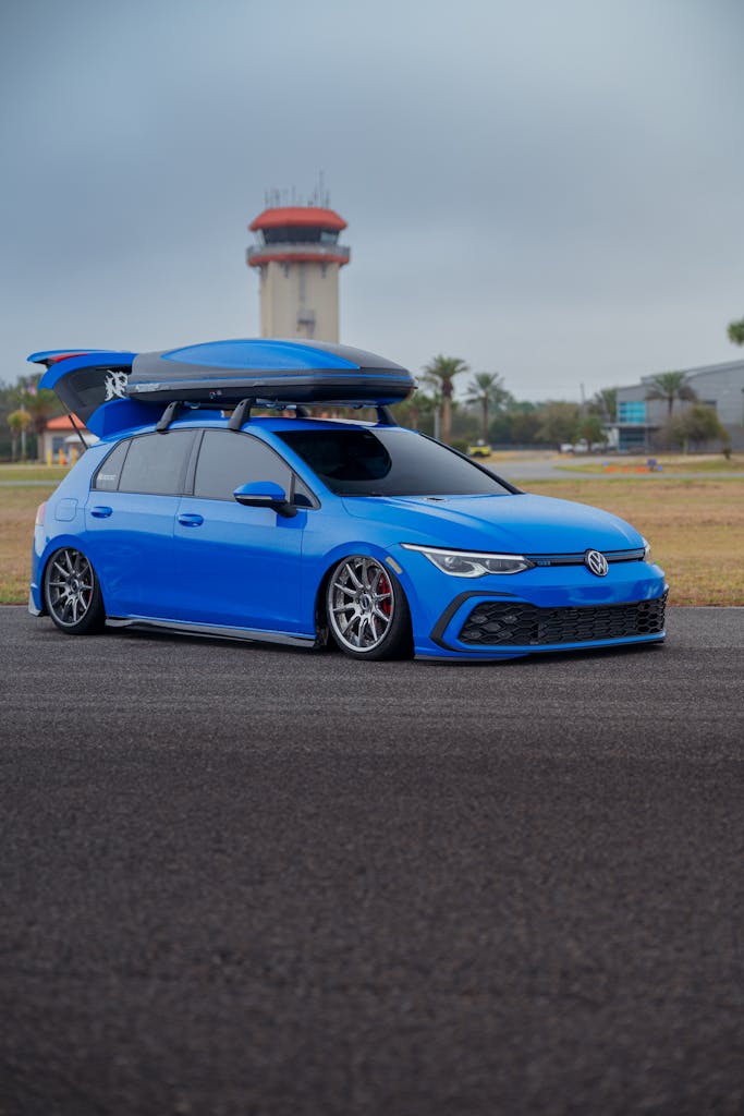 Airport Car Rental Qata A sleek blue hatchback car with a roof box parked near an airport with control tower in view.