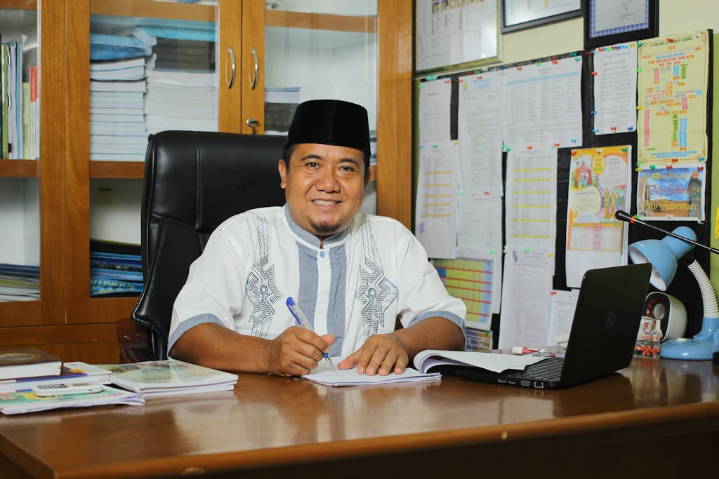 Dojen Moe An Indonesian teacher in traditional attire, smiling at his desk with documents in an office setting.