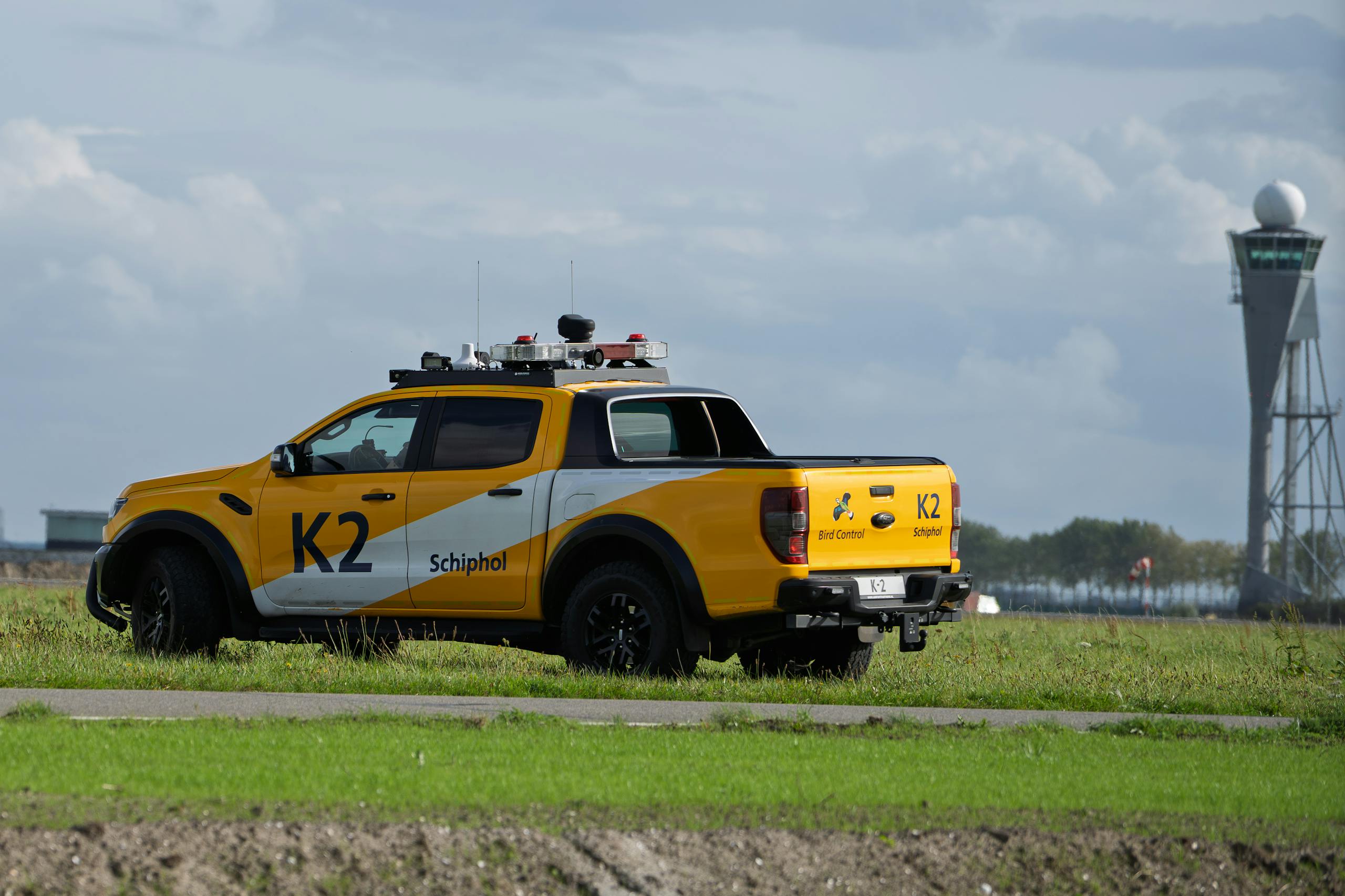 Airport Car Rental Qata Schiphol Bird Control vehicle parked on grass near airport tower under cloudy skies.
