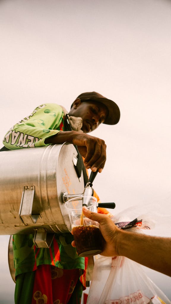 Matarecycler Street vendor pours a cold drink from a metal canister into a customer's cup on a warm day.