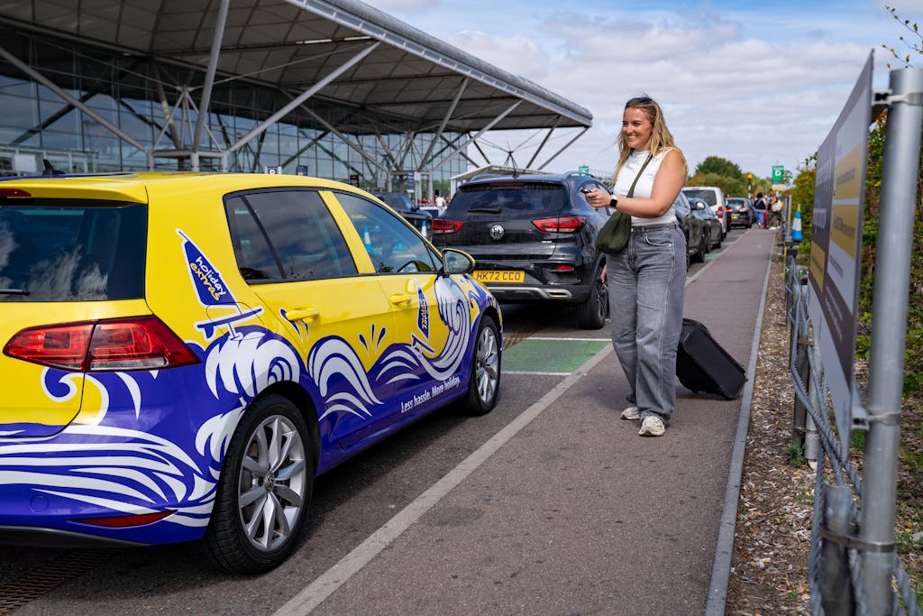 Airport Car Rental Qata Woman smiling by a colorful car at Stansted Airport's valet parking area.